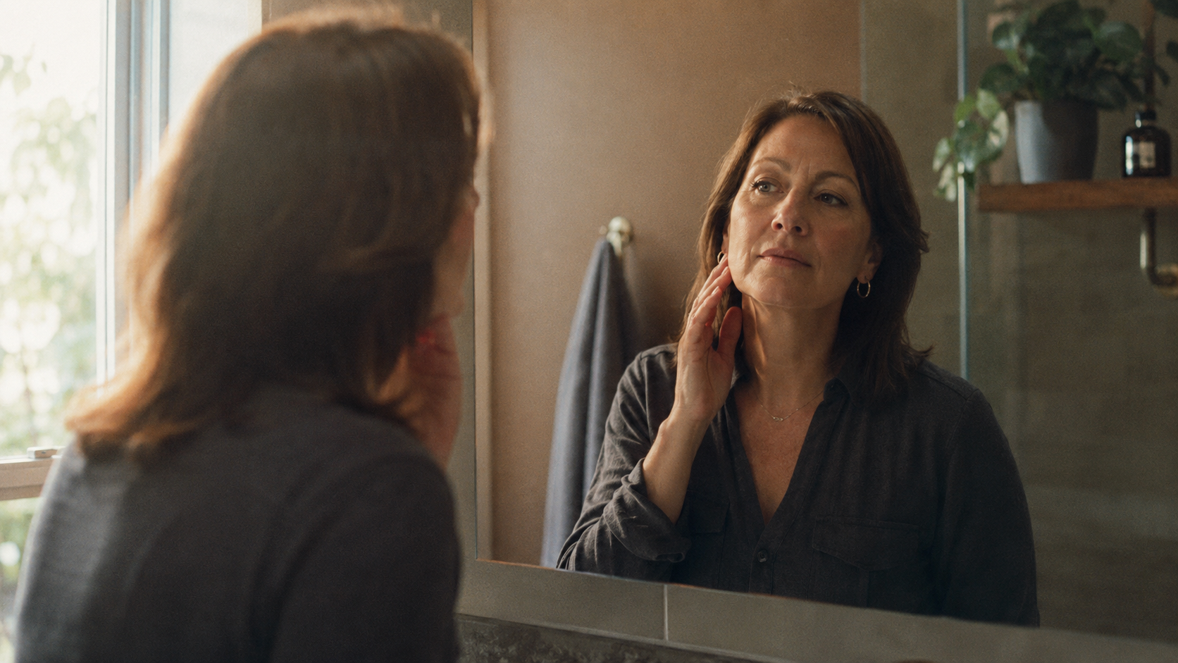 A woman in her mid-40s standing at a bathroom mirror with a thoughtful, composed expression — she's reached her weight loss goal and is calmly examining the subtle changes in her face.